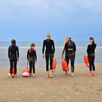 Groep kinderen in wetsuits op het strand met een instructeur en veiligheidsboeien