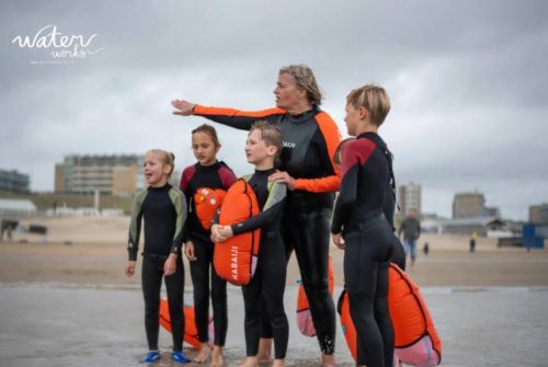 Groep kinderen in wetsuits op het strand met een instructeur en een veiligheidsboei